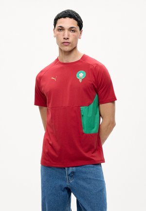 Jonge man met kort krullend haar, die een rood Puma T-shirt met een groen zijvak en het voetballogo van Marokko draagt, gecombineerd met een blauwe spijkerbroek.
