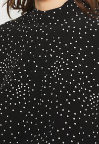Zwarte blouse met witte stippen, hoge kraag, subtiele plooien, zachte stof en een relaxte pasvorm. Patroon varieert in stipgrootte.