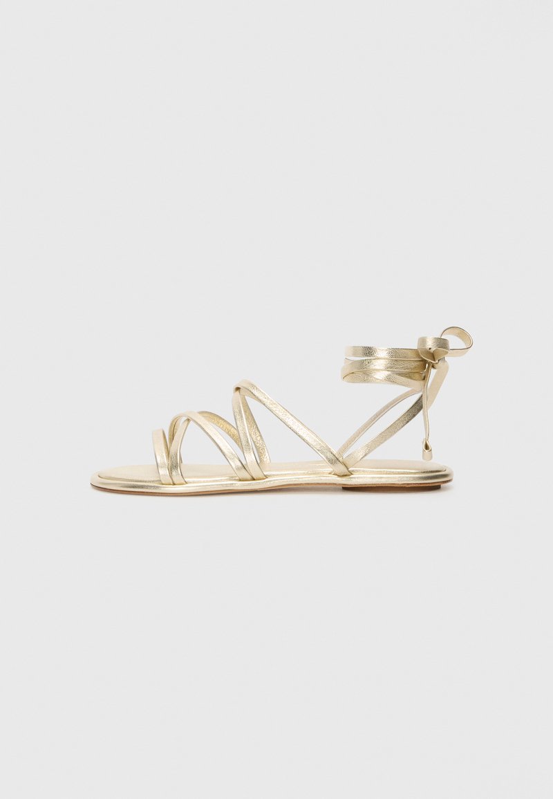 Gylne sandaler med stropper som har flat såle, flere tynne stropper og ankelinnpakning sikret med en sløyfe. Glatt metallisk tekstur.