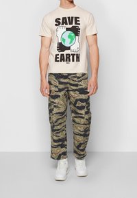 Muž oblečený v béžovém tričku "Zachraňte Zemi" s grafickým zobrazením rukou a globu, tigrími maskáčovými cargo kalhotami a bílými teniskami.