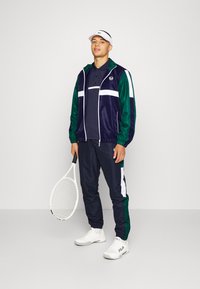 Námořnicky modrý a zelený sportovní trenčkot s bílými akcenty, zahrnující zipovou bundu, odpovídající kalhoty a polo tričko. Drží tenisovou raketu.