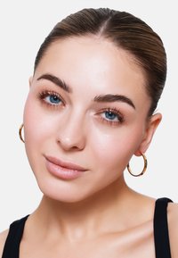 Close-up van het gezicht van een persoon met blauwe ogen, een zachte huid, lichte make-up en gouden hoepel oorbellen. Het haar is netjes naar achteren gekamd en de persoon draagt een zwarte top.