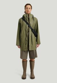 Oliven grøn vandtæt jakke med lynlås og trykknaplukning, parret med en matchende nederdel og brune gummistøvler. Sort crossbody taske bæres.