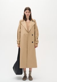 Beige trench coat with large lapels, double-breasted buttons, and oversized sleeves. Model holds a black tote bag, wearing brown socks and shoes.
