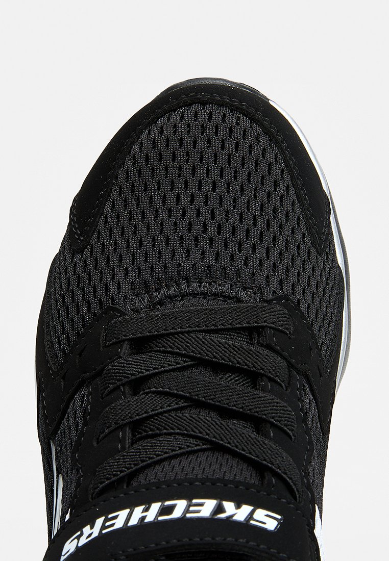 Zapatilla Skechers negra con parte superior de malla, cordones negros y logo blanco en la lengüeta, vista desde arriba.
