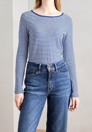 Personne portant un haut à manches longues rayé horizontalement bleu et blanc, rentré dans un jean en denim bleu taille haute avec double bouton devant.