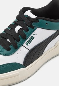 Sneakers PUMA con tomaia in pelle bianca, accenti neri e teal, pannello laterale testurizzato nero, lacci neri spessi e suola in gomma beige.