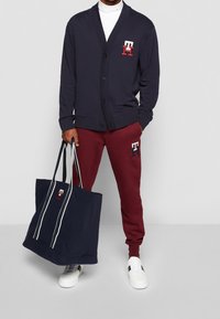 Cardigan blu navy con bottoni, dolcevita bianco, pantaloni jogger color borgogna con logo e una grande borsa a tracolla blu navy con strisce bianche e logo. Scarpe da ginnastica bianche.