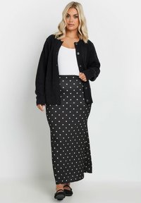 Mustikka harmaa neuleliivi, jossa on nappikiinnitys, käytetty valkoisen paidan ja mustan pilkkulakin maksi-hameen päällä. Hameessa on valkoisia pisteitä matalla pinnalla.