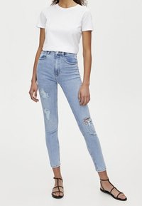 Jeans de denim de tiro alto, de color azul claro, con detalles desgastados y un ajuste ceñido. Combinados con una camiseta blanca sencilla y sandalias negras.