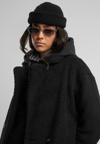 Black teddy coat with a textured, fluffy material, layered over a black hoodie. Black beanie and rectangular sunglasses complete the look.