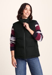 Smiling woman zipping black puffer vest, wearing striped purple, white, and maroon sweater and blue jeans, standing against beige background.