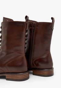 Botas de tobillo de cuero marrón con un acabado suave, cierres con cordones y cremallera, tacón apilado y un sutil detalle de costura en los costados.