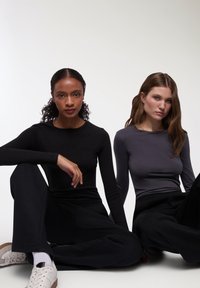 Due modelli seduti, che indossano magliette a maniche lunghe: uno in nero e l'altro in grigio scuro. Entrambi hanno pantaloni neri casual e sneakers bianche.