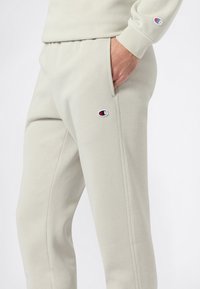 Pantalones de chándal gris claro fabricados en un tejido suave, con cinturilla elástica, bolsillos laterales y un pequeño parche de logo de color en el muslo izquierdo.