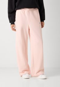 Pantalons de survêtement rose clair, à jambes larges, avec une taille élastique et des détails de couture discrets. Associés à des chaussures blanches.