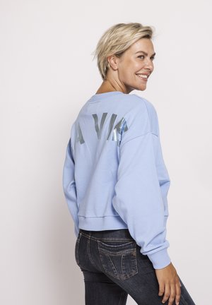 Smiling woman with short blonde hair wears light blue sweatshirt with silver "LA VIE" text, paired with dark jeans, turned slightly sideways.