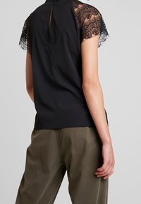 Top nero con maniche in pizzo a cappuccio, dettaglio a chiave sulla schiena e texture liscia. Abbinato a pantaloni verde oliva per un contrasto.