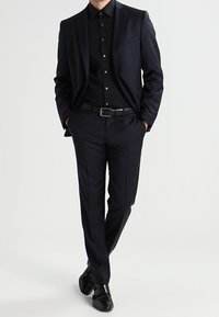 Dark tailored suit featuring a single-breasted jacket and slim trousers, accented by a black button-up shirt and polished shoes.