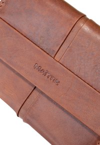 Portefeuille en cuir marron avec logo "MAÎTRE" en relief, présentant une texture lisse et une forme rectangulaire avec coutures et plis visibles.