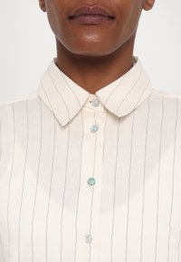 Chemise à col en tissu écru avec de fines rayures verticales. Détails remarquables : boutons turquoise et texture lisse.