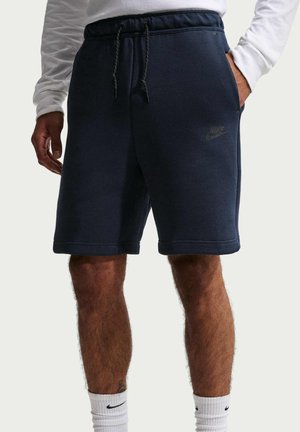 Man draagt marineblauwe Nike-short met trekkoord, wit shirt met lange mouwen en witte Nike-sokken, één hand in de zak, staat rechtop.