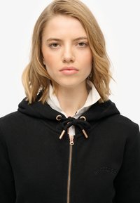 Felpa nera con zip e cappuccio regolabile, dotata di hardware in tono dorato e logo in rilievo sul petto. Materiale in cotone, texture morbida.