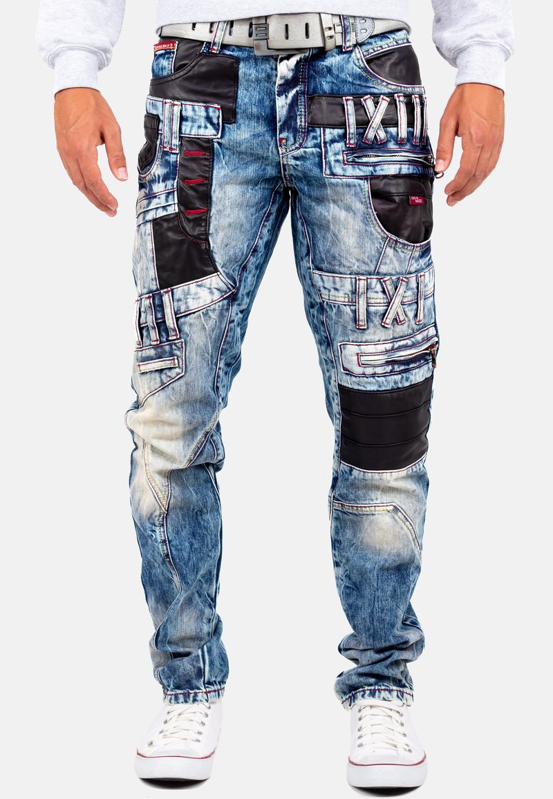 Pantalons cargo en denim bleu avec un design à poches multiples, ornés de détails en cuir noir, de coutures rouges et d'une finition légèrement déchirée.