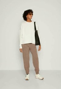 Suéter blanco oversized, pantalones jogger marrones, zapatillas blancas y un bolso tote negro. Modelo con cabello rizado y gafas de sol se encuentra de pie contra un fondo liso.