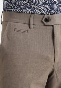 Närbild av beige byxor med svart knappstängning, bältesöglor, smal sidficka och en marinblå mönstrad skjorta instoppad.