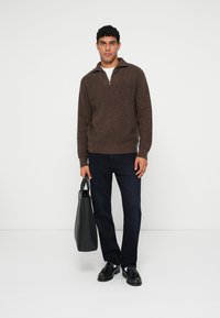 Brun, strikket, lynlås-sweater kombineret med mørke bukser og sorte sko. Modellen holder en sort tote bag. Simpelt design, tekstureret stof.