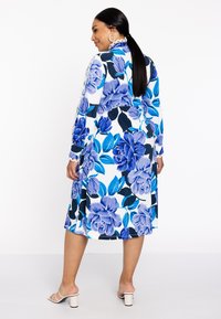 Bloemenjurk in het wit, met grote blauwe rozen en groene bladeren, met een middellange snit en knoopdetails op de mouwen.