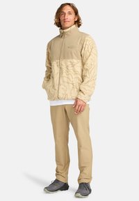 Veste en polaire beige avec un motif texturé, col montant et fermeture éclair, associée à un pantalon beige et des chaussures de randonnée grises.