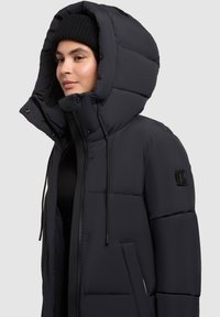 Donna che indossa un cappello di lana nera e una giacca a vento nera con zip e grande cappuccio, in piedi contro uno sfondo grigio chiaro uniforme.