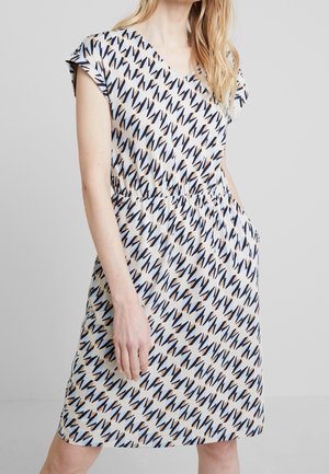 Femme portant une robe à manches courtes avec un motif géométrique zébré noir et beige et une taille élastique, debout devant un fond uni.
