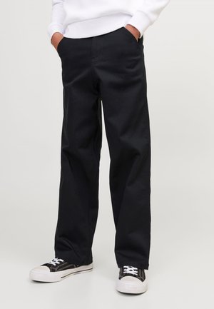 Personne portant un pantalon noir ample et des baskets noir et blanc, debout les mains dans les poches devant un fond clair uni.