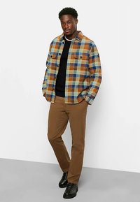 Kariertes Hemd in Orange-, Blau- und Gelbtönen, mit Knöpfen und zwei Brusttaschen. Schwarzer Pullover darunter, kombiniert mit hellbraunen Hosen und schwarzen Schuhen.