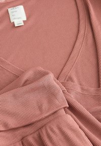 Mjukt rosa ribbat tyg med V-ringad design och slät textur. Inkluderar en etikett med texten "NEXT time to dream" sydd i halsringningen.