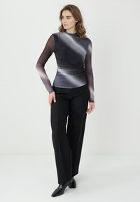 Kobieta stoi, ubrana w dopasowaną czarno-szarą bluzkę z długim rękawem o gradientowym wzorze, szerokie czarne spodnie oraz czarne, szpiczaste buty, na tle jednolitej białej ściany.