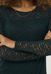 Dark green lace top featuring long sleeves with a textured pattern. Two delicate gold necklaces complement the design.