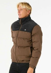 Bruine pufferjack met een zwarte corduroy schouderpartij, ritssluiting, elastische manchetten en zijzakken. Gelaagd materiaal met een gestoeld ontwerp.