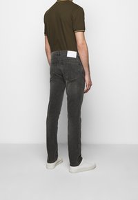 Osoba w ciemnoszarych dżinsach, oliwkowej koszulce polo z krótkim rękawem i białych sneakersach, stojąca na tle gładkiej białej ściany.