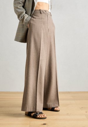 Pantalones marrones de pierna ancha combinados con sandalias negras, un top blanco corto y un blazer de cuadros sobre un fondo de suelo de madera clara.