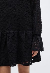 Μαύρο δαντελένιο φόρεμα με λουλουδένια μοτίβα, βολάν στο τελείωμα και μανίκια καμπάνα. Το ύφασμα έχει υφή με διάφανα στοιχεία.