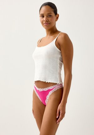 Weißes, geripptes Tanktop mit gewelltem Saum, kombiniert mit einem rosa Tanga mit Spitzenbesatz und einer Schleifendetail vorne.