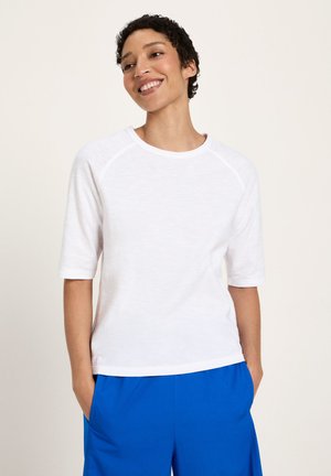 Lächende Person mit kurzen lockigen Haaren, die ein weißes Halbarmhemd und blaue Hosen trägt, Hände in den Taschen, vor einfachem Hintergrund.