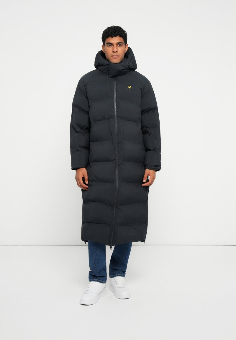 Abrigo acolchado negro largo con un cuello alto, cremallera frontal y diseño acolchado. Lleva puesto sobre unos vaqueros azules y zapatillas blancas.