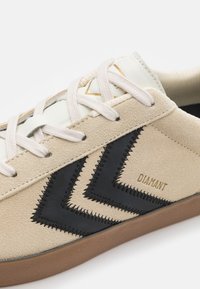 Beżowe zamszowe sneakersy z czarnym zygzakowym przeszyciem, białymi skórzanymi akcentami i teksturowanymi sznurówkami, na brązowej gumowej podeszwie. Złoty detal z logo.