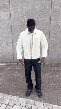 Light cream zip-up jacket with side pockets, worn over a light gray shirt. Black cargo pants with multiple side pockets. Black shoes.