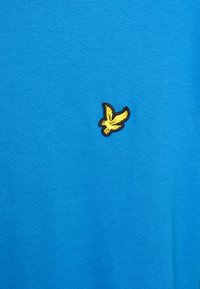 Tricou din bumbac albastru, cu un mic logo brodat în formă de pasăre galbenă pe partea stângă a pieptului. Textură fină, design casual.